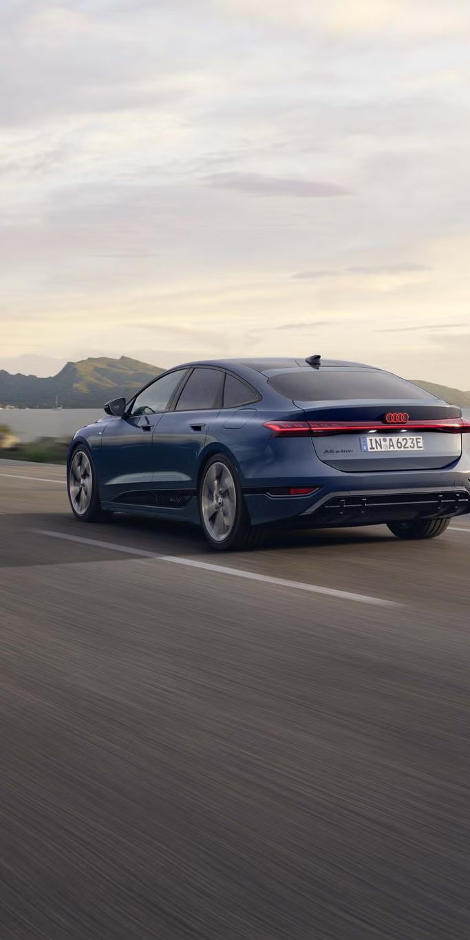 Der A6 Sportback e-tron aus schräger Heckansicht fährt eine Straße parallel zum Ufer entlang. Im Hintergrund sind Berge zu erkennen. 