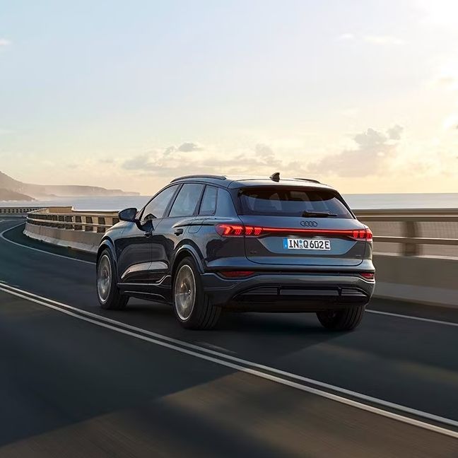 Pohľad na zadné partie Audi Q6 počas dynamickej jazdy po ceste popri pobreží.