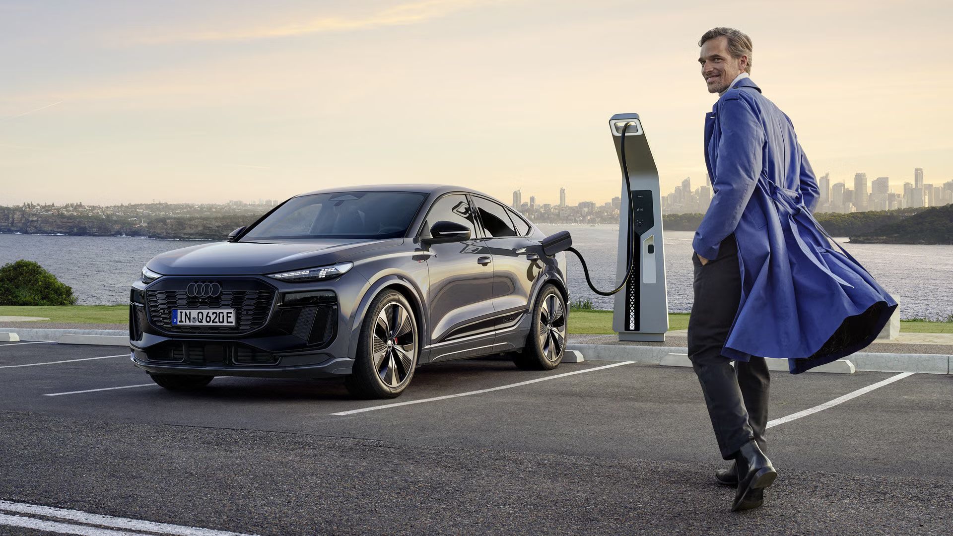 Audi Q6 e-tron je zaparkovaný na parkovisku pri verejnej nabíjacej stanici. Elegantný muž prichádza k vozidlu, obzerá sa cez rameno a pozerá do objektívu.