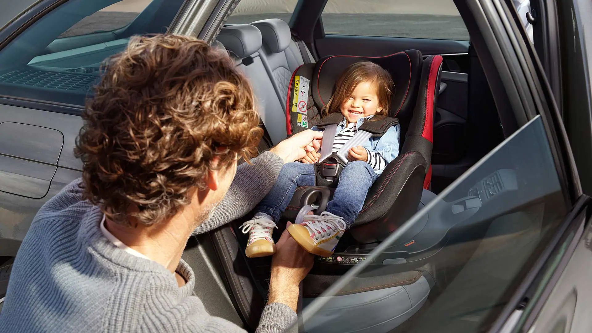 Muž upevňuje bezpečnostný pás malej dieťaťu sediacemu v autosedačke. Autosedačka je umiestnená na zadných sedadlách v aute. Dvere auta sú otvorené.
