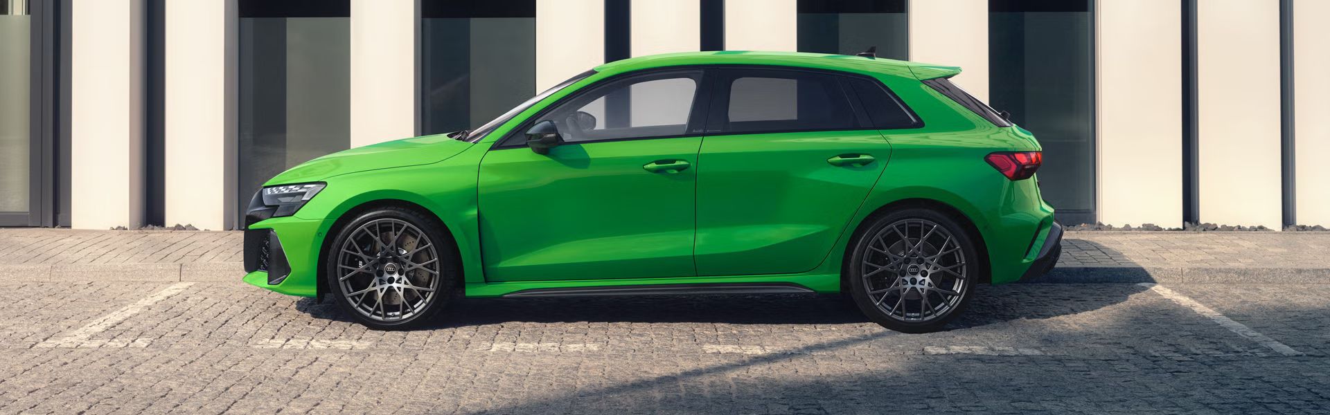 RS 3 Sportback zaparkovaný na dlažobných kockách pri chodníku vedľa verejnej, modernej budovy.