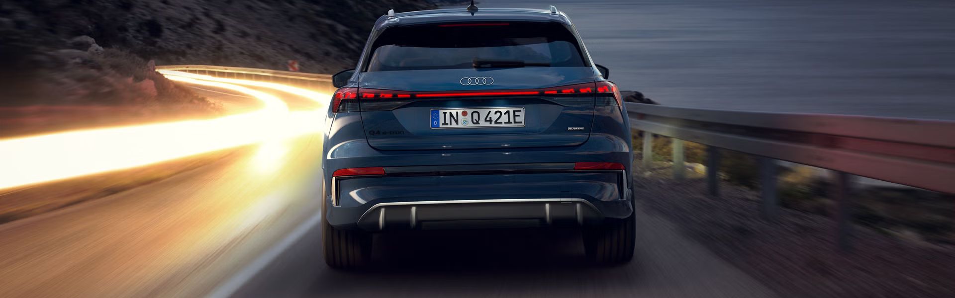 Audi Q4 SUV e-tron zozadu v noci na strmej horskej ceste, vpravo je more. V protismere sú vidieť svetelné línie prechádzajúcich vozidiel. 