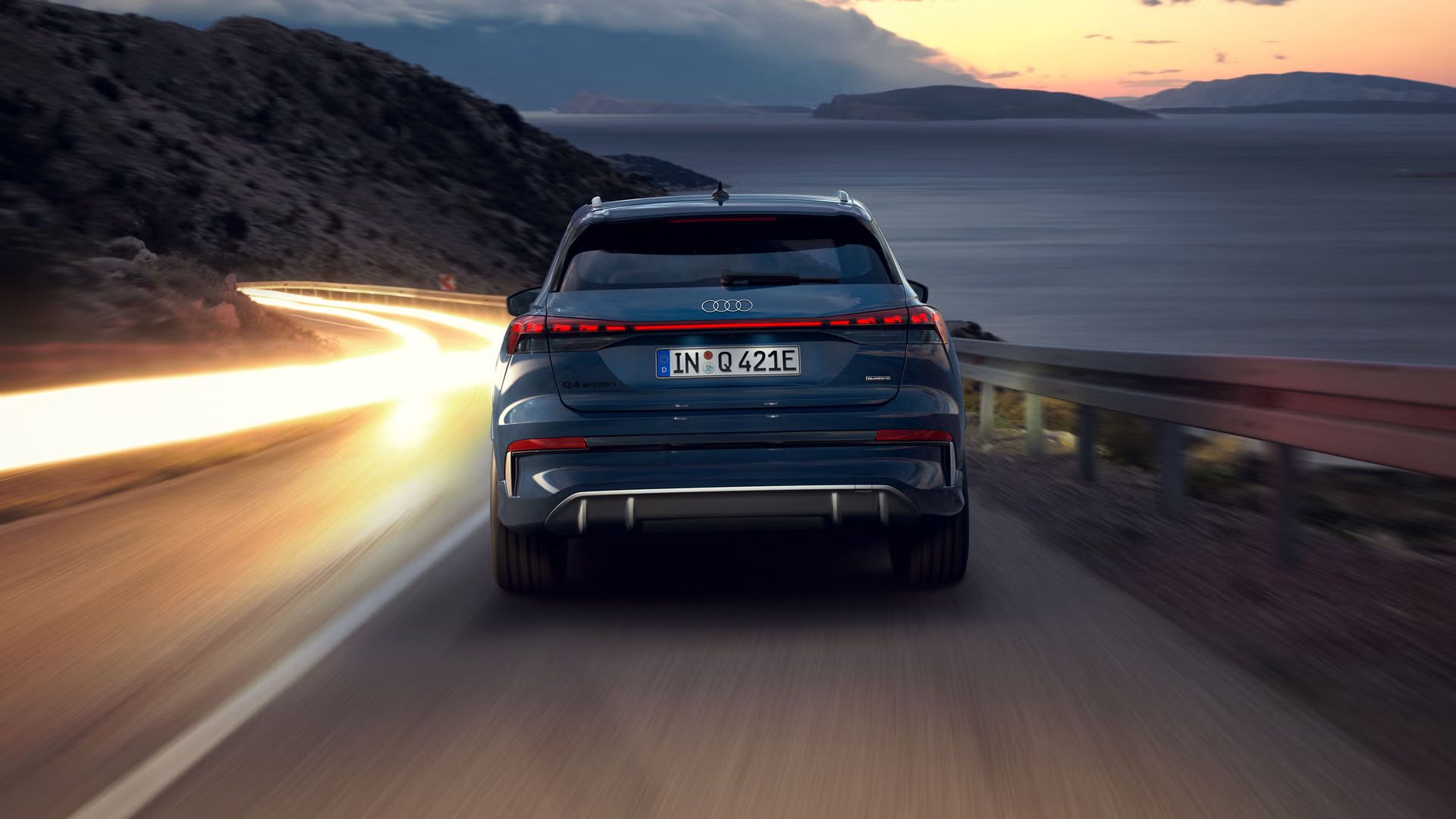 Audi Q4 SUV e-tron zozadu v noci na strmej horskej ceste, vpravo je more. V protismere sú vidieť svetelné línie prechádzajúcich vozidiel. 