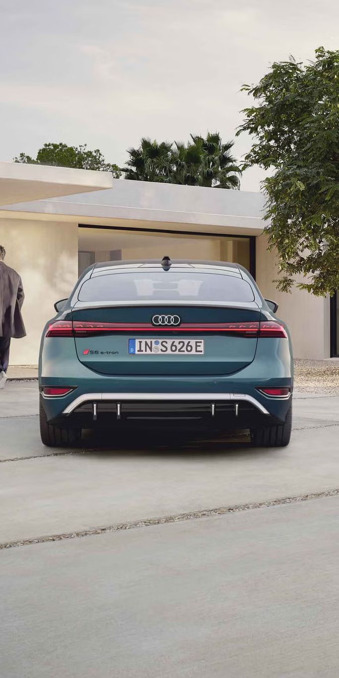 Der S6 Sportback e-tron im Heckprofil. Das Fahrzeug ist dem Eingangsbereich eines modernen Gebäudes zugewandt. Vor dem Auto befindet sich ein junger Mann, der auf das Gebäude zuläuft.