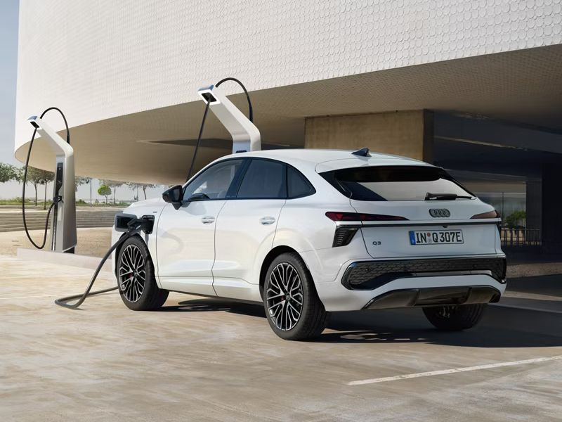 Audi Q3 Sportback e-hybrid zaparkované pred svetlou verejnou budovou so zaujímavou fasádou pri verejnej nabíjacej stanici. Vozidlo sa nabíja. V pozadí vidieť schody, stromy, zeleň a oblohu.