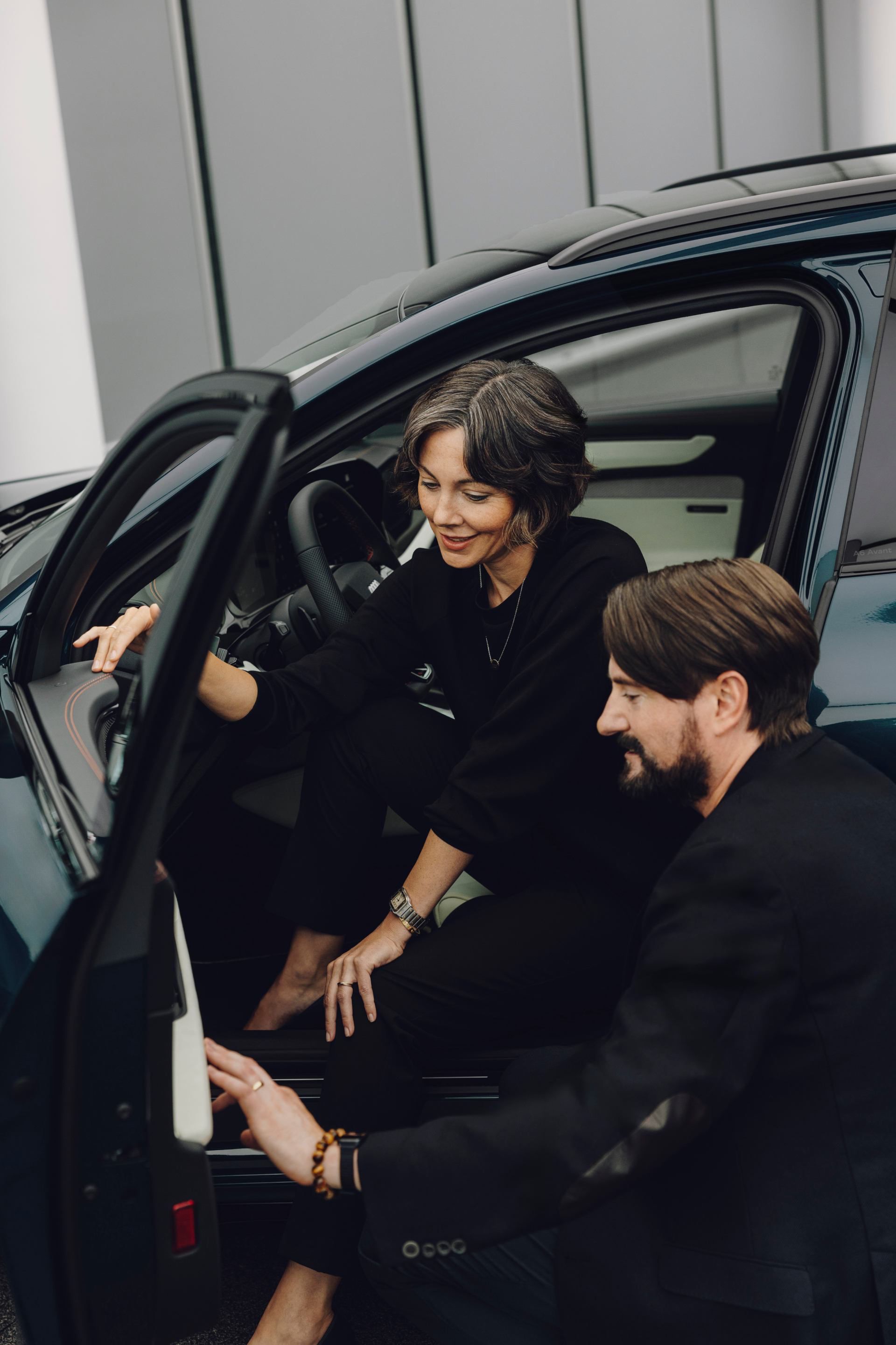 Dve osoby sú pri aute. Jedna osoba sedí vo vnútri auta a druhá čupí vedľa otvorených dverí. Obe sú oblečené v tmavom oblečení.