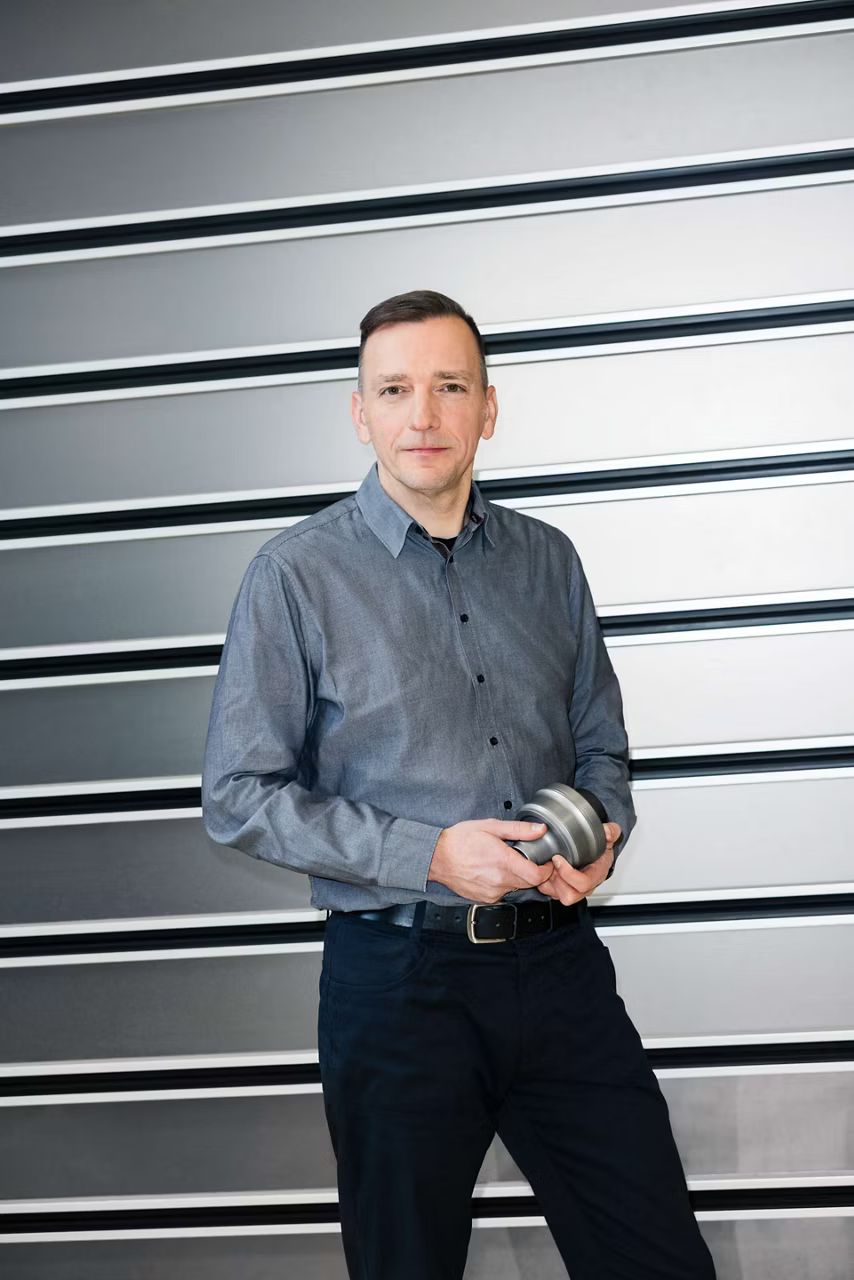 Muž stojí pred sťahovacou bránou, pozerá sa do kamery a v rukách drží súčiastku.