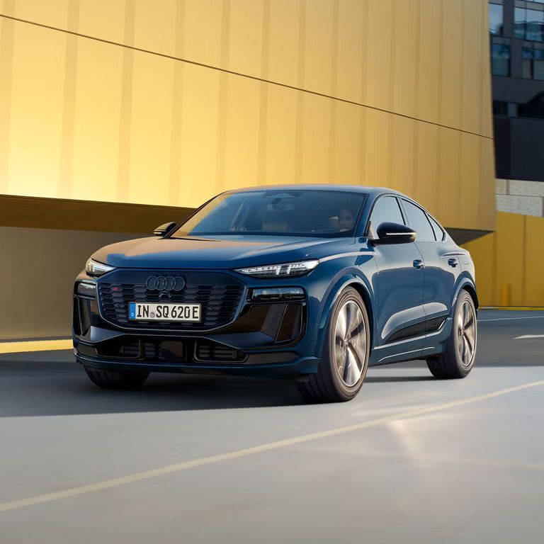 Audi SQ6 Sportback e-tron jazdí po ceste v meste. Za vozidlom je vidieť verejnú budovu so žltou fasádou.