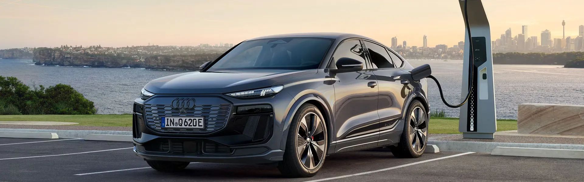 Moderné čierne elektrické Audi Q6 Sportback je zaparkované na parkovisku pri nabíjacej stanici pri mori. Nabíjačka je pripojená k autu a disky kolies majú športový dizajn. V pozadí je mestská panoráma so zapadajúcim slnkom.