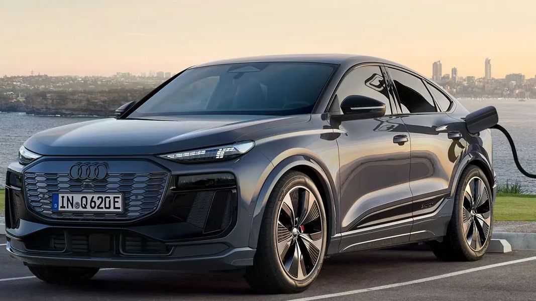 Moderné čierne elektrické Audi Q6 Sportback je zaparkované na parkovisku pri nabíjacej stanici pri mori. Nabíjačka je pripojená k autu a disky kolies majú športový dizajn. V pozadí je mestská panoráma so zapadajúcim slnkom.
