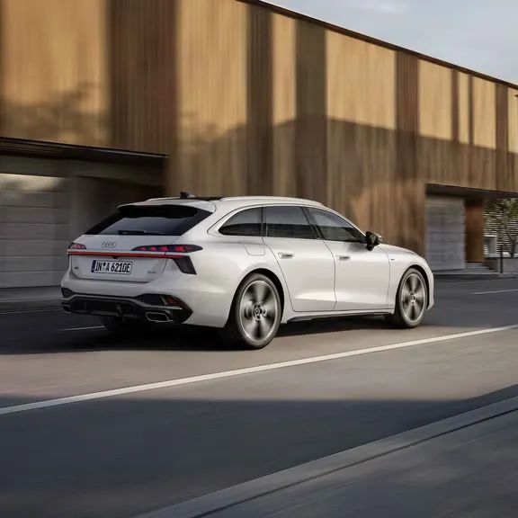 Audi A6 Avant e-hybrid počas jazdy v meste. Vozidlo prechádza okolo budovy s drevenou fasádou.