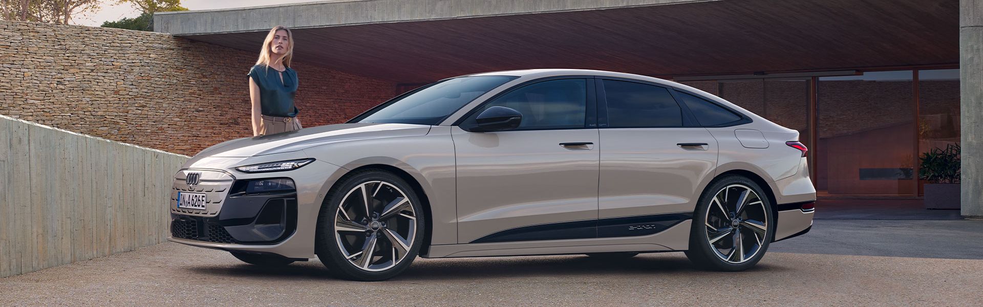 Elektrické Audi A6 Sportback e-tron zaparkované pred moderným domom pri západe slnka. Vľavo pri vozidle stojí mladá žena.