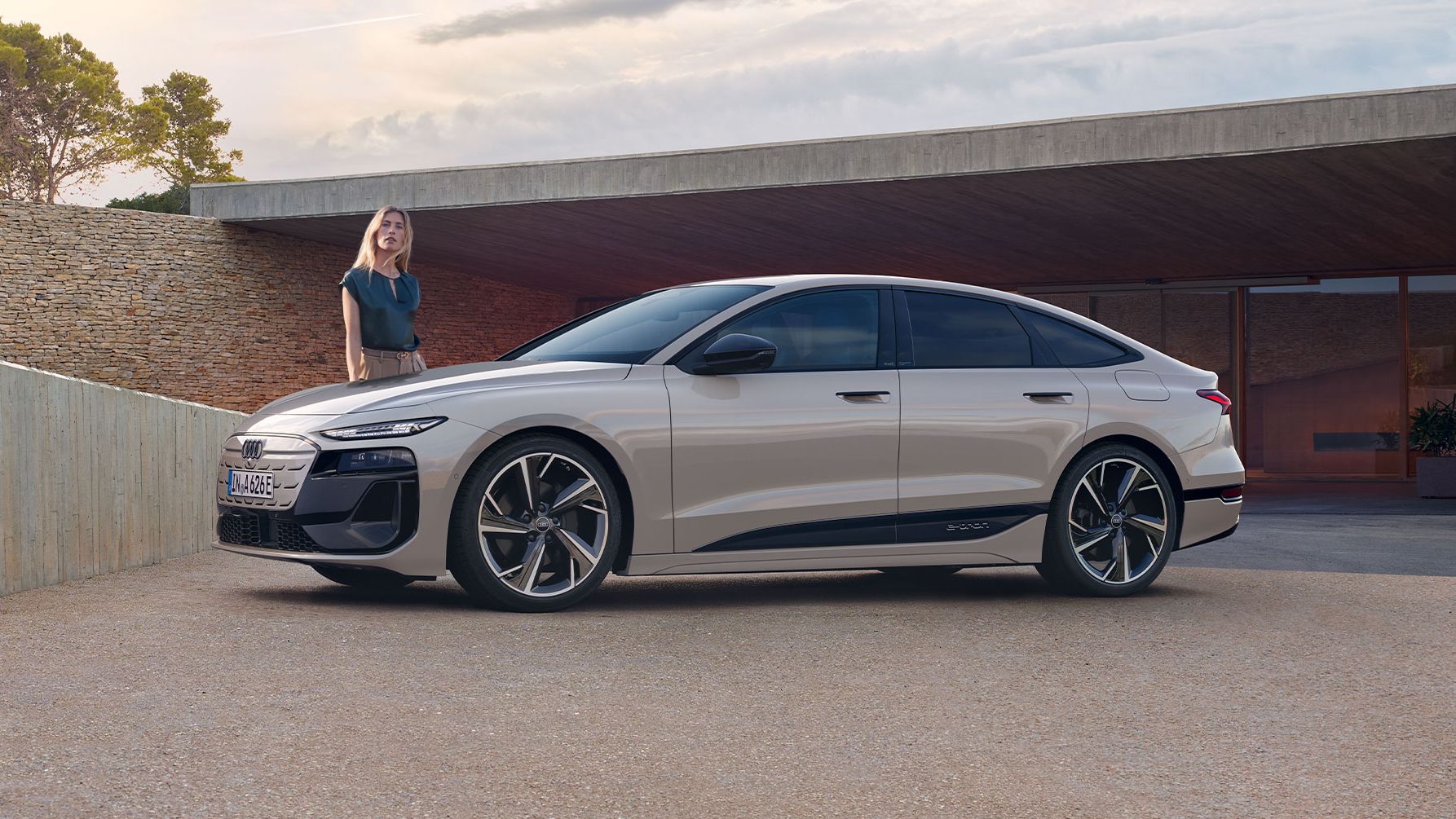 Elektrické Audi A6 Sportback e-tron zaparkované pred moderným domom pri západe slnka. Vľavo pri vozidle stojí mladá žena.