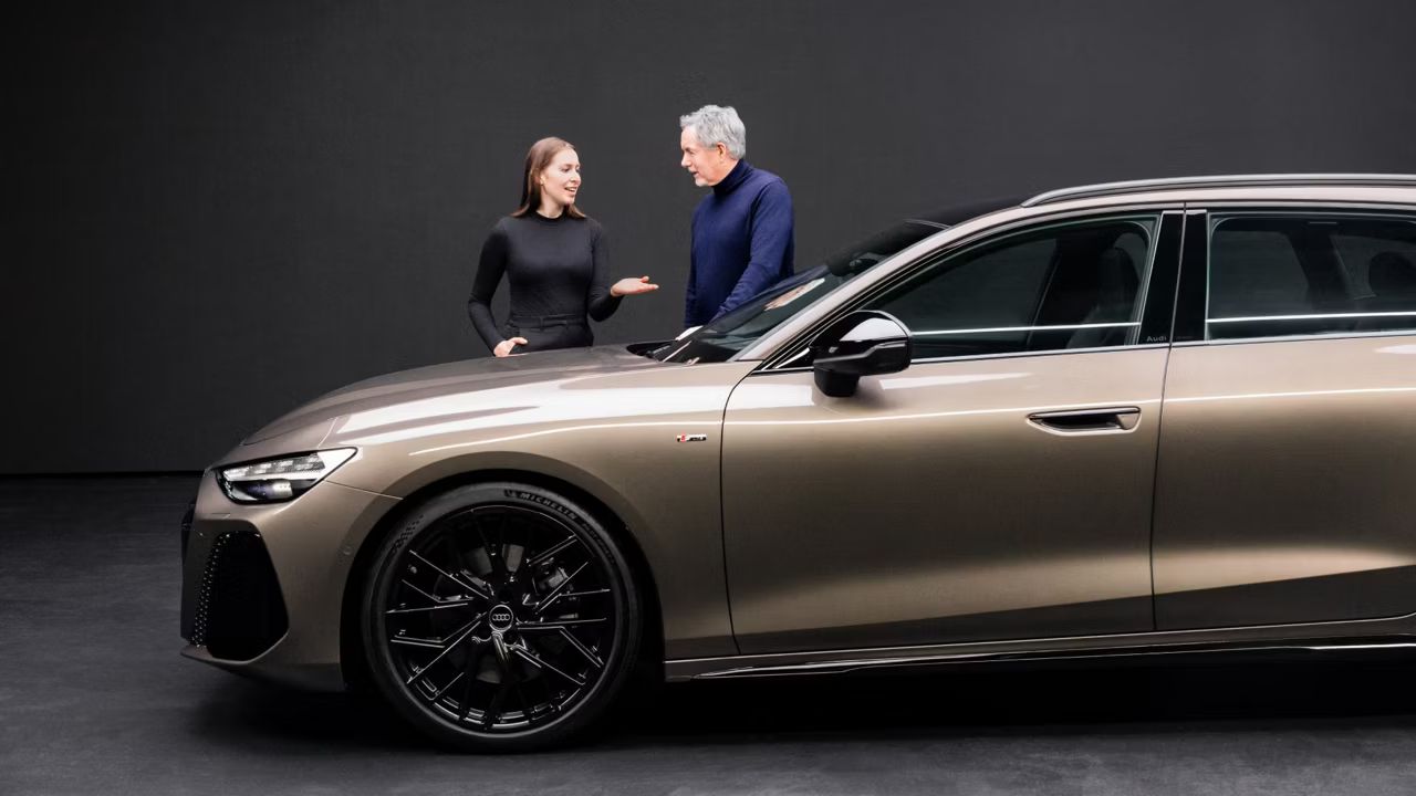 Audi A6 Avant v hnedej metalíze zaparkované v prezentačnom priestore pred tmavoantracitovou stenou. Za vozidlom stojí muž a žena a diskutujú spolu.