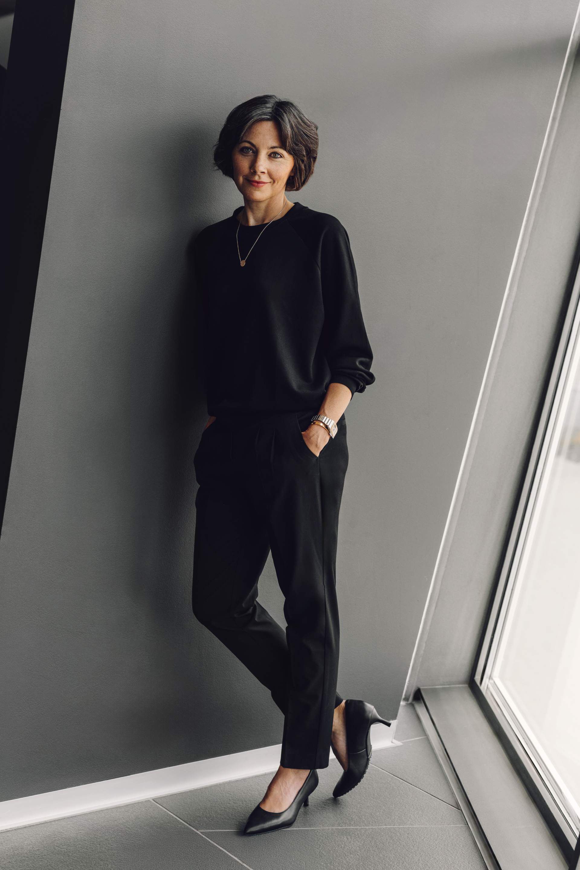 Žena v čiernom elegantnom oblečení stojí opretá o stenu. Má na sebe čierny top, nohavice a lodičky. Je v interiéri vedľa veľkého okna.
