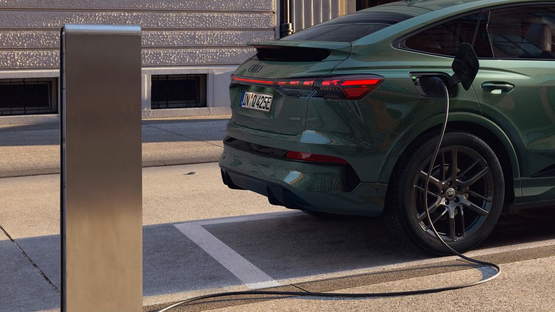 Pohľad na pravú zadnú časť vozidla Q4 Sportback e-tron, zaparkovaného pri nabíjacej stanici. V pozadí je moderná budova. Kábel nabíjacej stanice je pripojený k vozidlu.