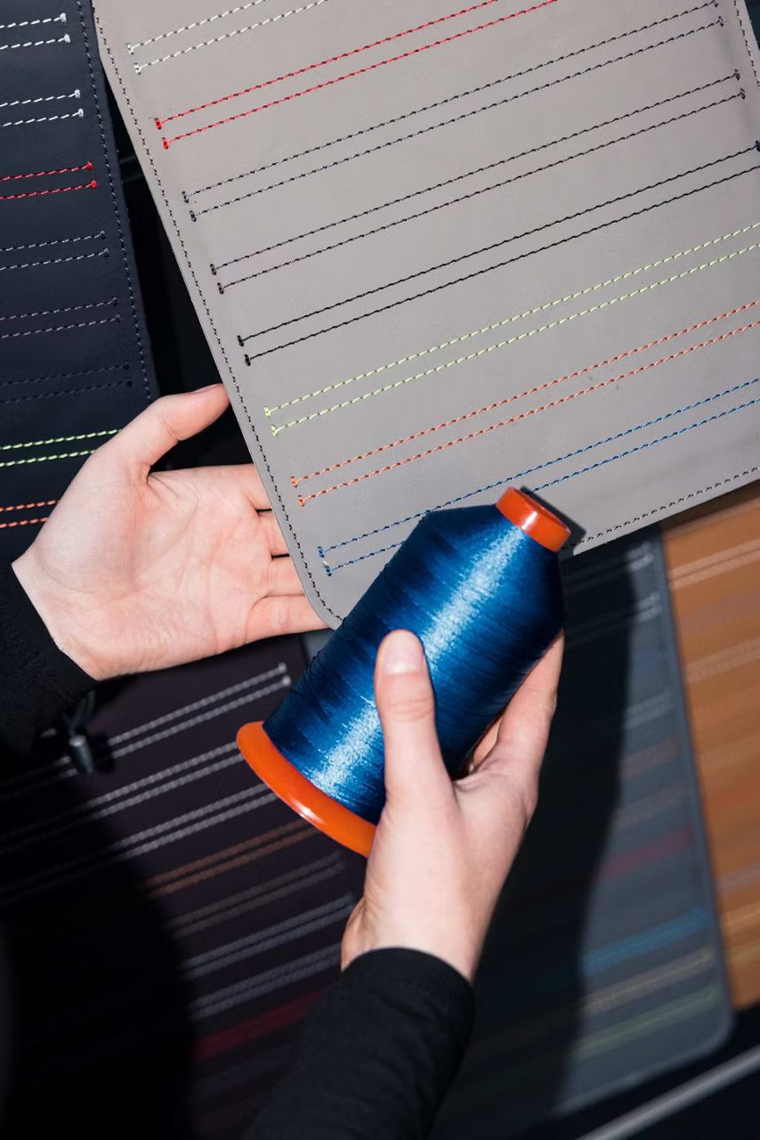 Detail na kus hnedosivej kože s rôzne farebným prešívaním. Vedľa nej je rovnaký kus kože v tmavomodrom odtieni. Vidíme tiež dve ruky, jedna drží sivohnedú kožu , druhá cievku modrej nite.