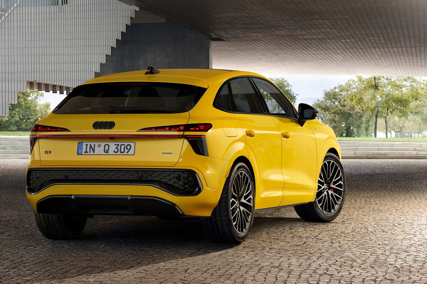 Žlté Audi Q3 Sportback je zaparkovaný na dláždenej ploche vedľa schodiska.
