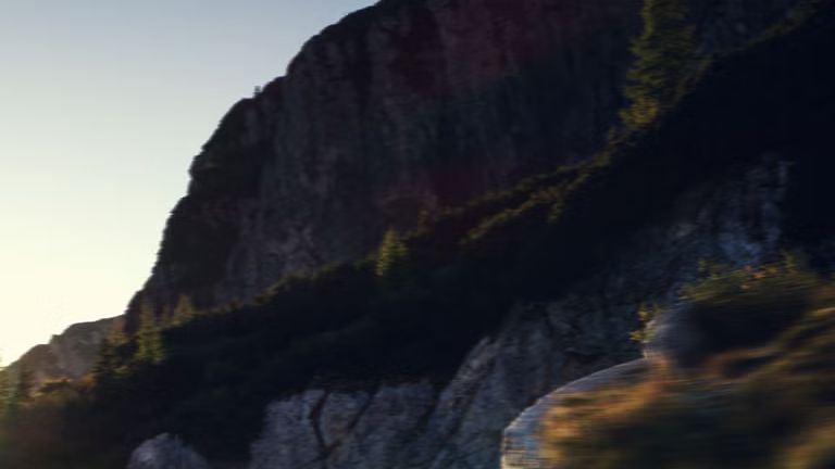 Q4 Sportback e-tron počas jazdy po horskej ceste. Slnečné lúče zľava hore osvetľujú obraz, vozidlo vrhá tieň dopredu.
