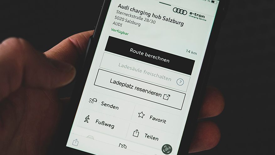 Ruka drží mobilný telefón s otvorenou aplikáciou na vyhľadávanie nabíjacích staníc pre elektrické vozidlá. Na obrazovke sú viditeľné možnosti ako vypočítať trasu, rezervovať nabíjacie miesto a zdieľať informácie. Zobrazená je aj poloha konkrétnej nabíjacej stanice Audi v Salzburgu.