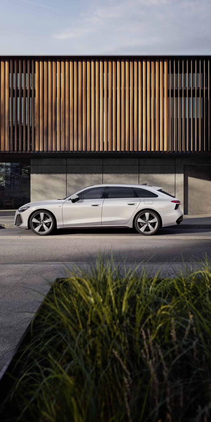 Der A6 Avant e-hybrid steht im Seitenprofil in einer Parkbucht vor der Fassade eines Gebäudes.