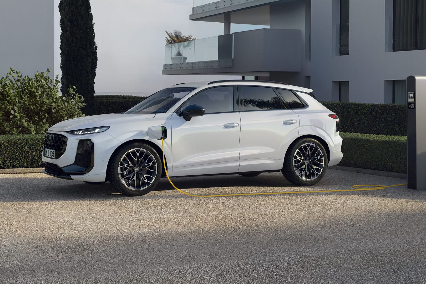 Biele Audi Q3 SUV e-hybrid zaparkované na parkovisku pri obytných domoch. Vozidlo je pripojené k verejnej nabíjacej stanici a nabíja sa.