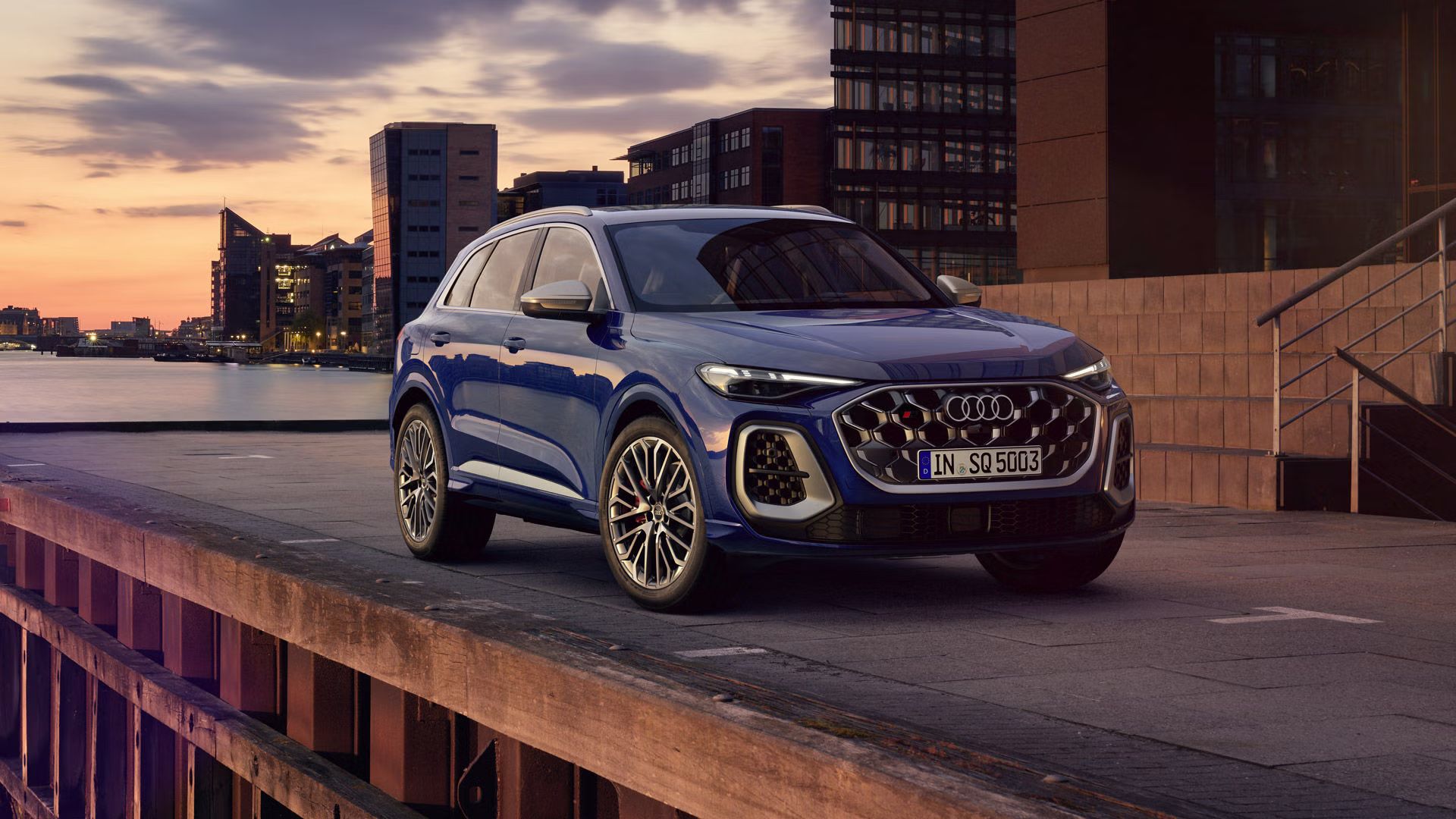 SUV SQ5 stojí pred panorámou pobrežného mesta. Pred ním stojí mladá, módna žena. 