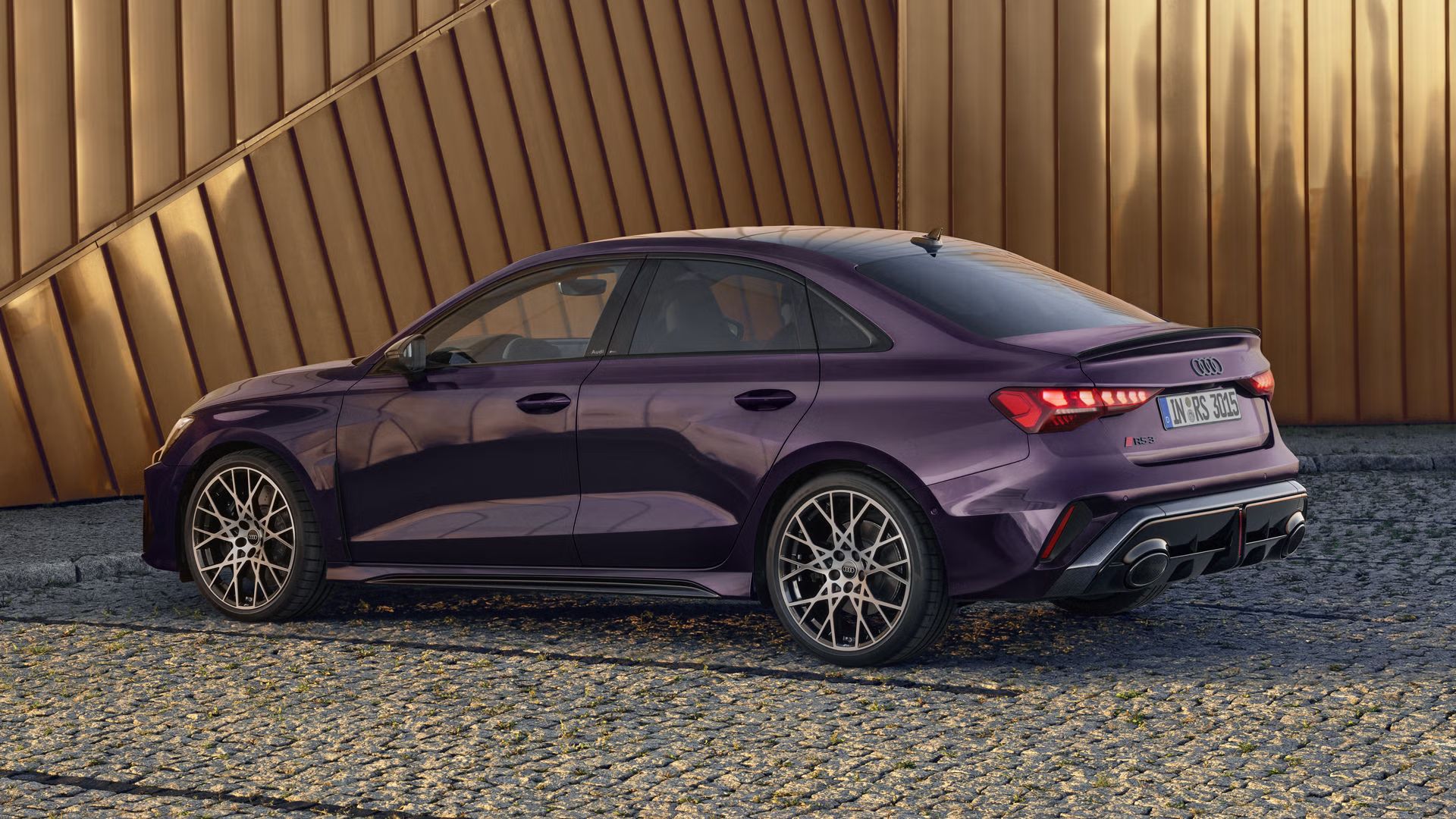 Audi RS 3 Limuzína s individuálnym lakovaním v odtieni Merlin s perleťovým efektom zaparkovaný na dlažobných kockách, v pozadí vidieť obrysy modernej budovy.