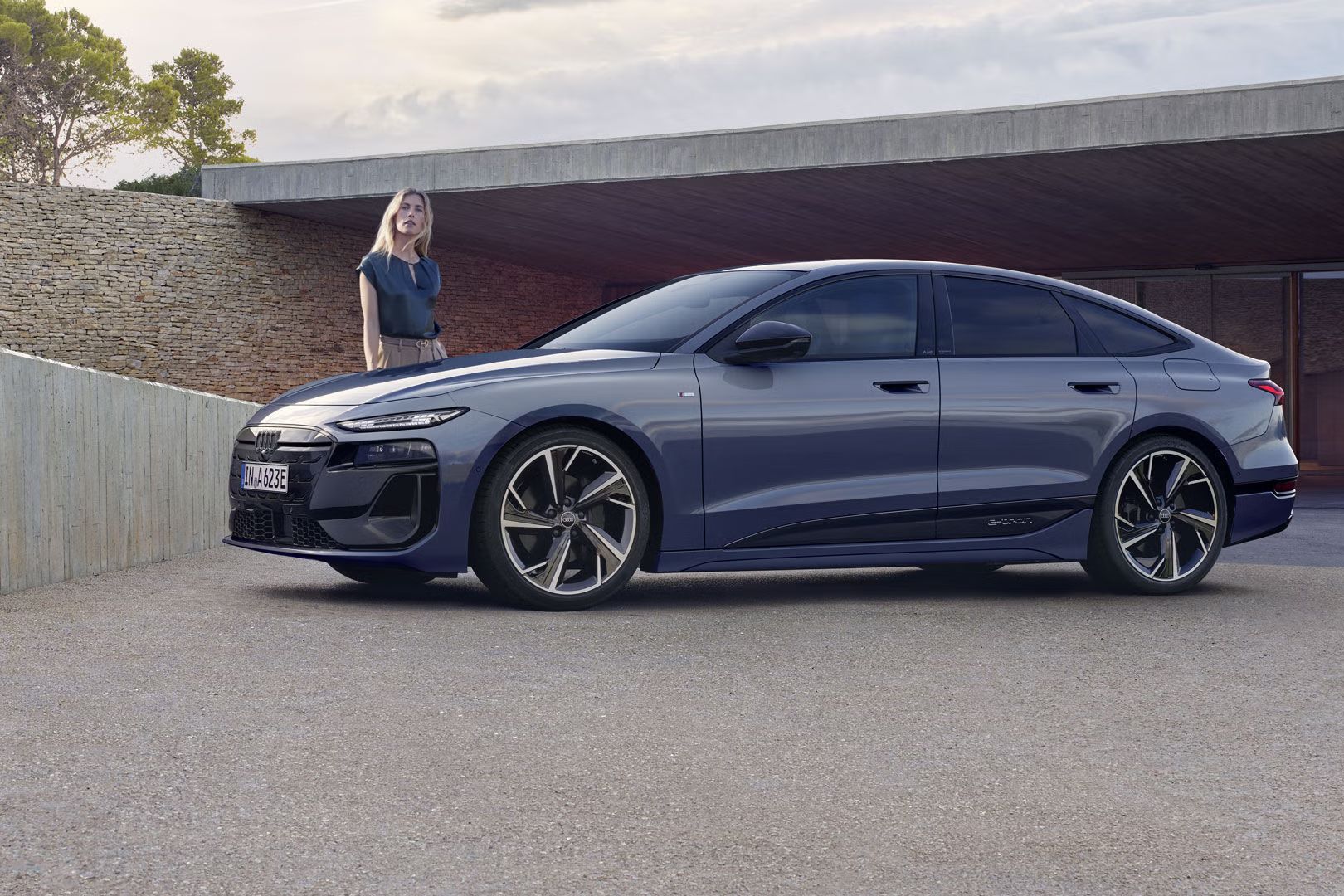 A6 Sportback e-tron stojí v bočnom profile pred garážou domu. Vedľa neho stojí mladá žena.