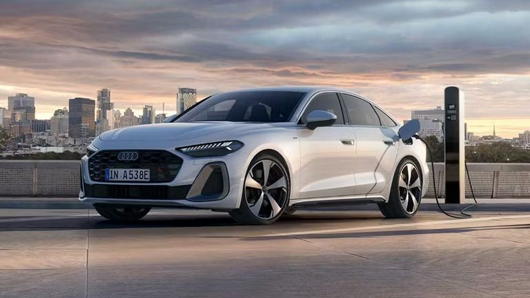Strieborné Audi A5 Limuzína e-hybrid stojí na parkovisku vedľa nabíjacej stanici a nabíja sa. V pozadí je mesto pri zatiahnutej oblohe a zapadajúcom slnku.