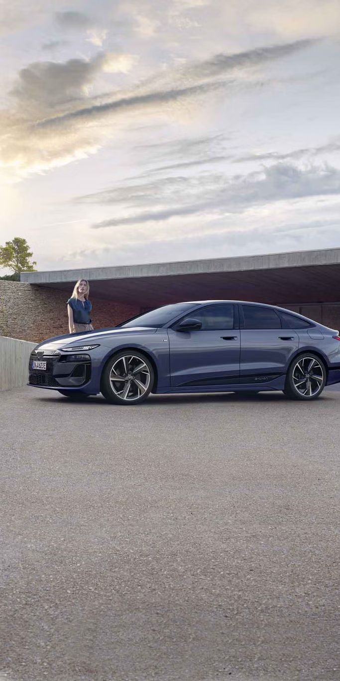 Der A6 Sportback e-tron steht im Seitenprofil vor dem Carport eines Hauses. Daneben steht eine junge Frau.