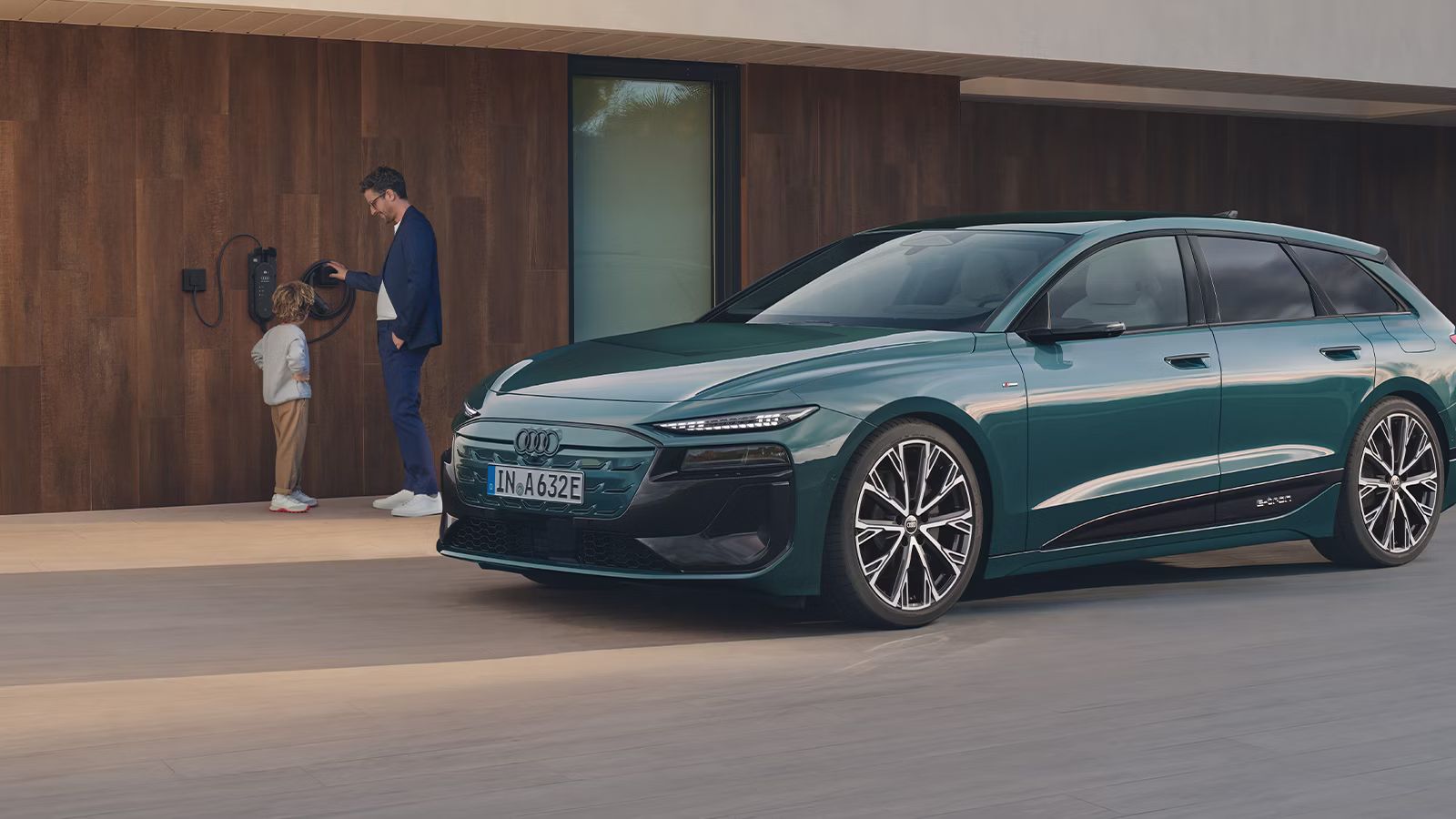 zelené Audi A6 Avant e-tron zaparkovaný pred moderným domom. Na fasáde domu je integrovaná domáca nabíjacia stanica. Muž so synom stoja pri tejto nabíjacej stanici. 