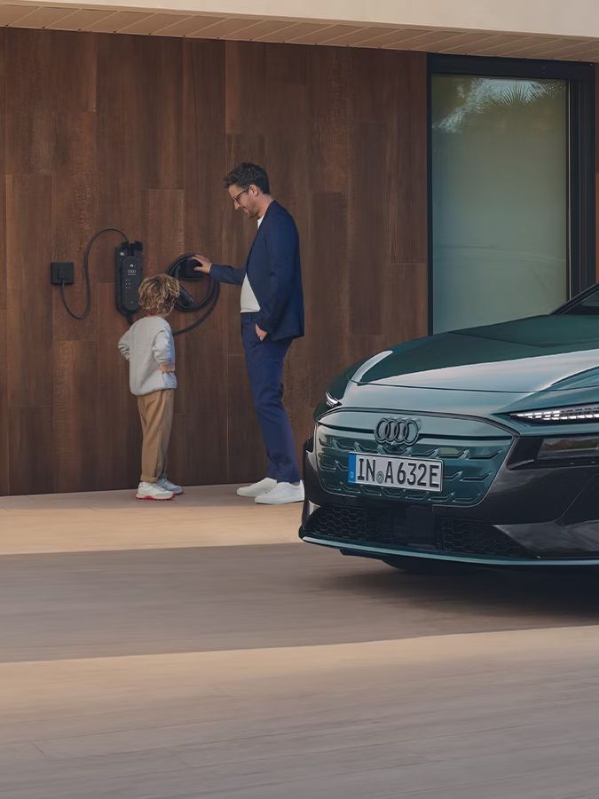 zelené Audi A6 Avant e-tron zaparkovaný pred moderným domom. Na fasáde domu je integrovaná domáca nabíjacia stanica. Muž so synom stoja pri tejto nabíjacej stanici. 