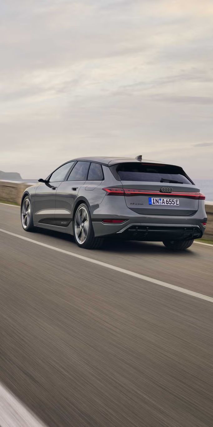 Schräge Heckaufnahme des A6 Avant e-tron, welcher auf einer Straße entlang des Meeres fährt. Im Hintergrund sind Felsen und Berge zu erkennen. 