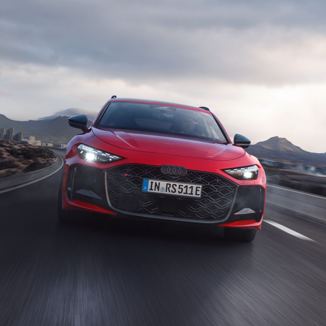 Červené Audi RS 5 Avant jazdí po ceste v horskej krajine. Obloha je zamračená.