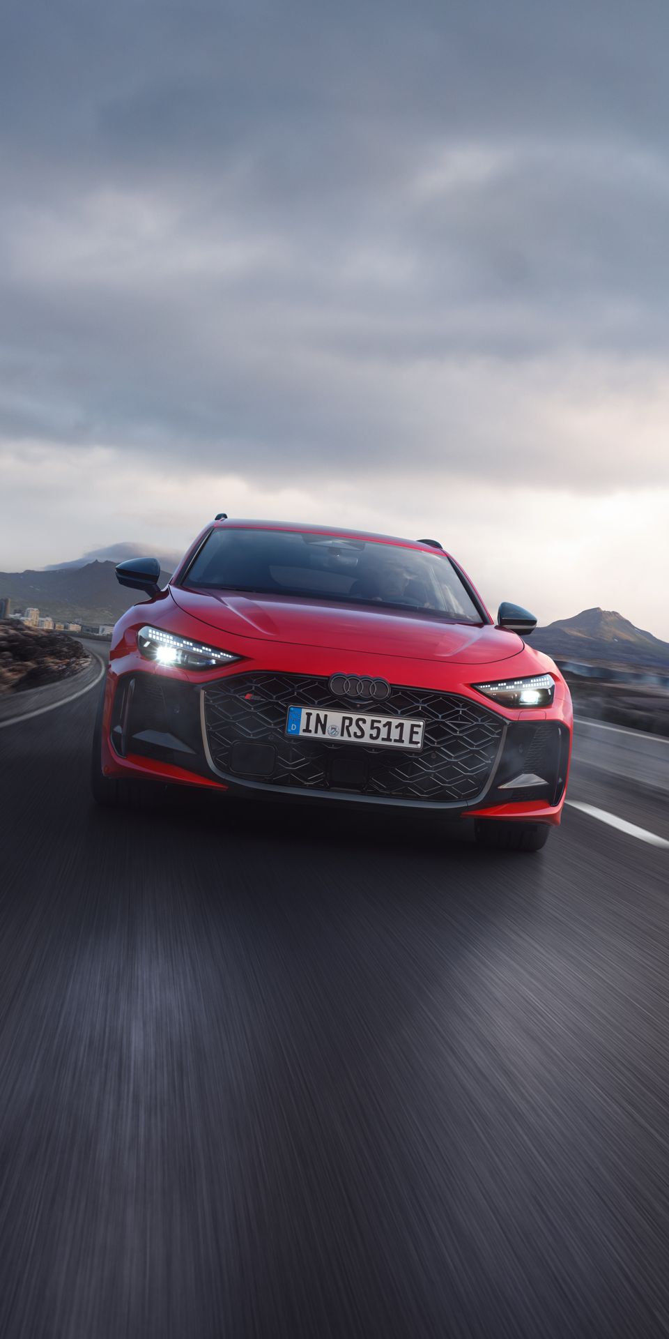 Červené Audi RS 5 Avant jazdí po ceste v horskej krajine. Obloha je zamračená.