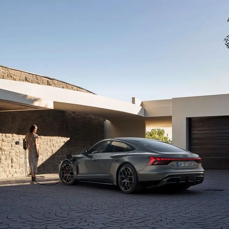 Audi RS e-tron GT zaparkované na dláždenej ploche pri modernej vile je napojené na domácu nabíjaciu stanicu. Vedľa vozidla stojí elegantne oblečená mladá žena.