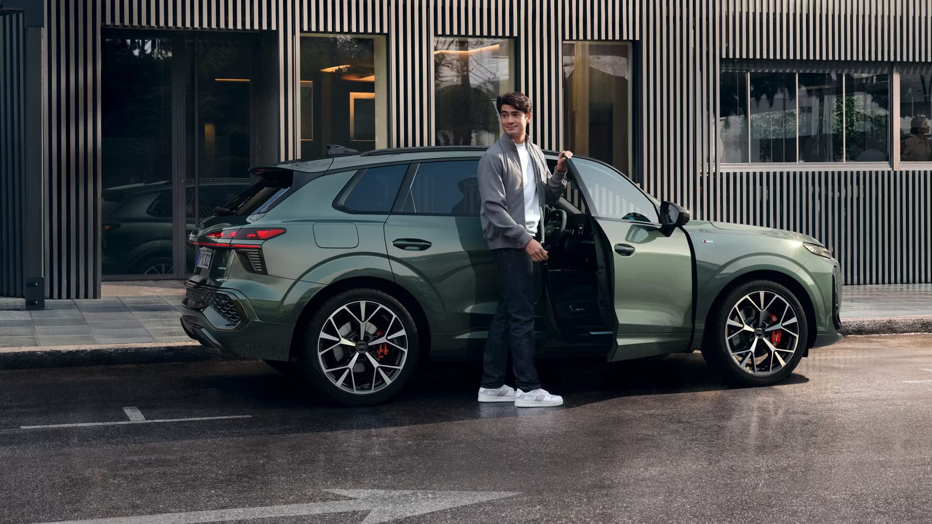 Audi Q3 SUV stojí na parkovacom mieste na ceste pri chodníku vedľa modernej mestskej budovy. Mladý muž drží otvorené dvere spolujazdca a pozerá sa cez rameno dozadu.