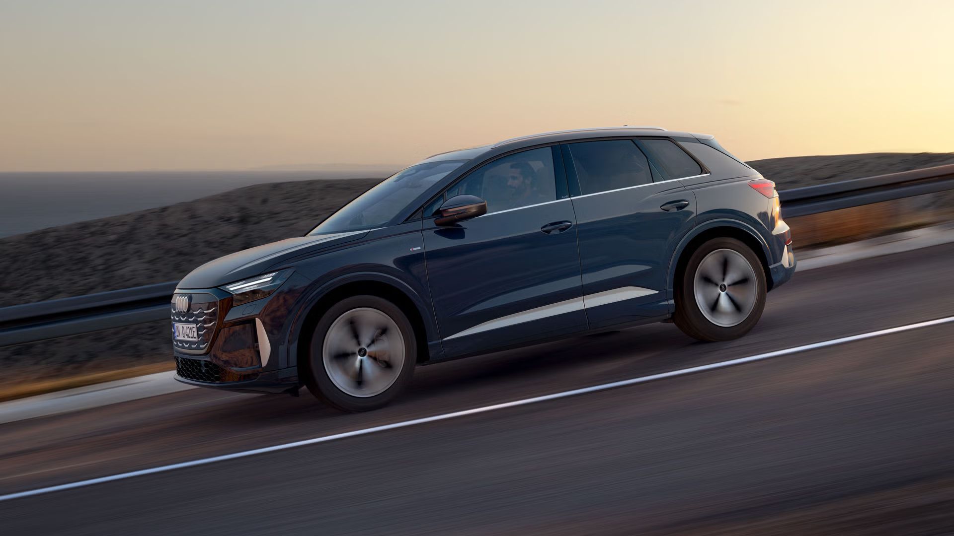 Pohľad zboku na Q4 SUV e-tron na klesajúcej ceste. V pozadí je vidieť more.
