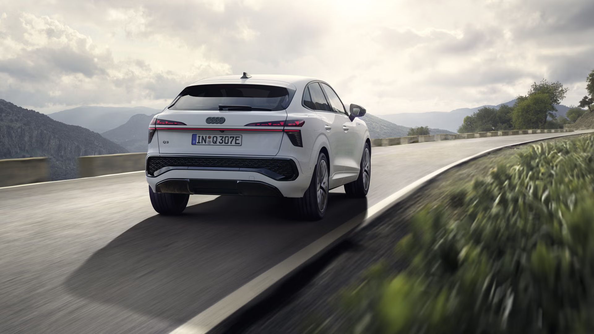 Biele Audi Q3 Sportback e-hybrid jazdí po vidieckej ceste v horskej krajine.