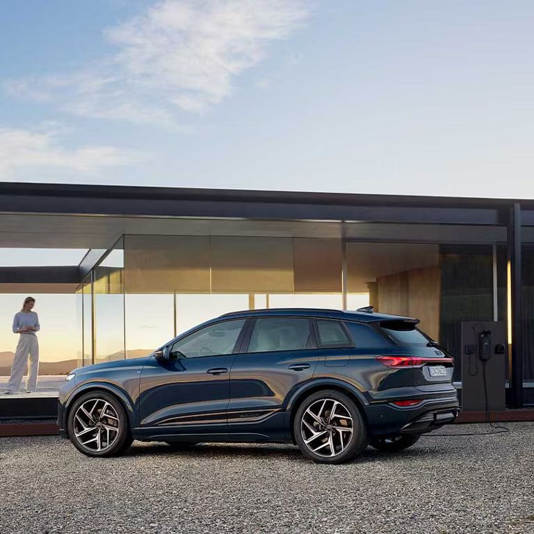 Audi Q6 SUV e-tron z bočného pohľadu. Zaparkovaný pred modernou vilou so sklenenou fasádou.