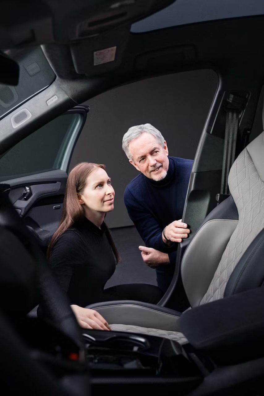 Muž a žena sa pozerajú do interiéru Audi A6 Avant vo výbave Audi exclusive v odtieni modrá Bajkala a neodýmová zlatá. Vidieť možno časť predného sedadla čalúneného kožou, bočné dvere, bezpečnostný pás, A stĺpik a časť čelného skla.