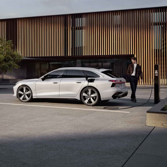 Der A6 Avant e-hybrid steht seitlich vor dem Eingangsbereich eines großen Gebäudes und wird über ein Stromkabel geladen. Am Heck des Fahrzeugs steht ein Mann mittleren Alters.
