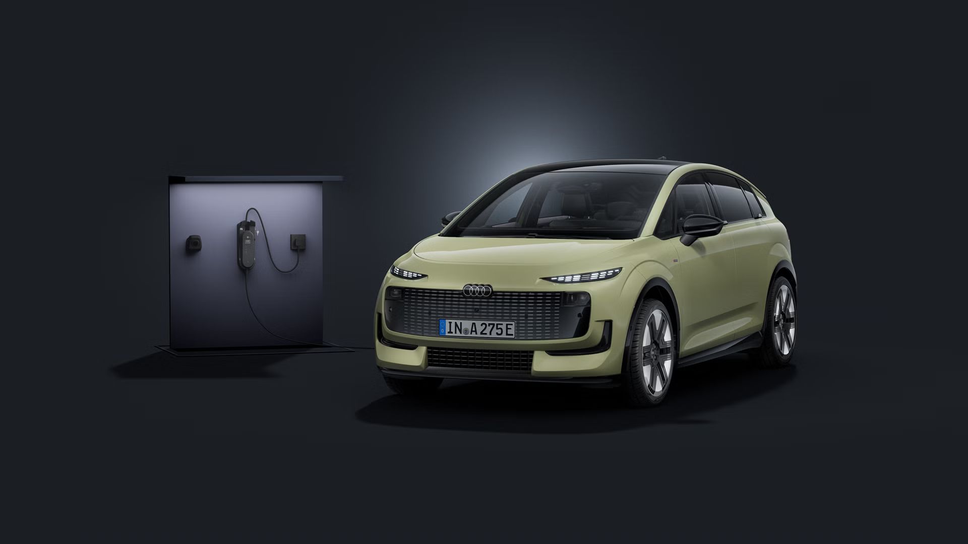 Audi A2 e-tron je šikmo spredu na tmavom pozadí, s nabíjacou stanicou vľavo za ním.