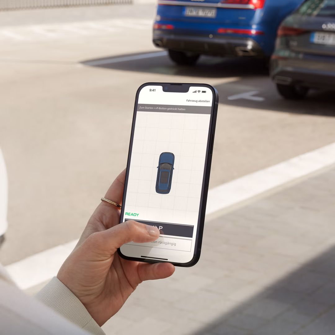 Módne oblečená mladá žena drží v ruke smartfón, na ktorom je vidieť štýlizované Audi z pohľadu zhora. Stojí pred zaparkovanými vozidlami.