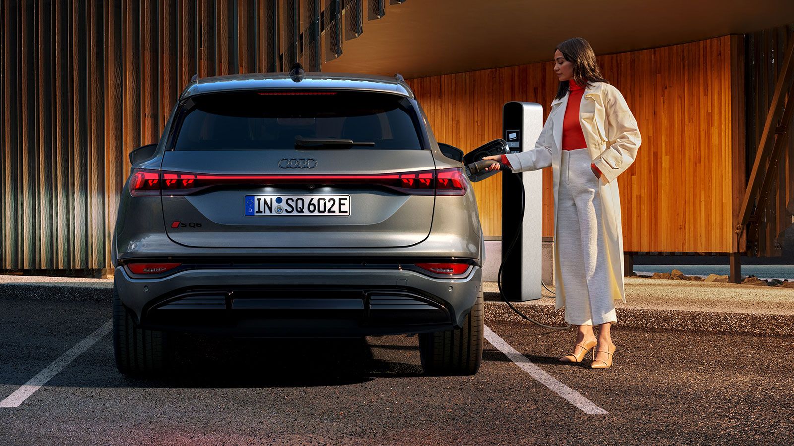 Audi SQ6 e-tron stojí na parkovacom mieste a je pripojený k nabíjacej stanici. Mladá žena drží nabíjací konektor. 