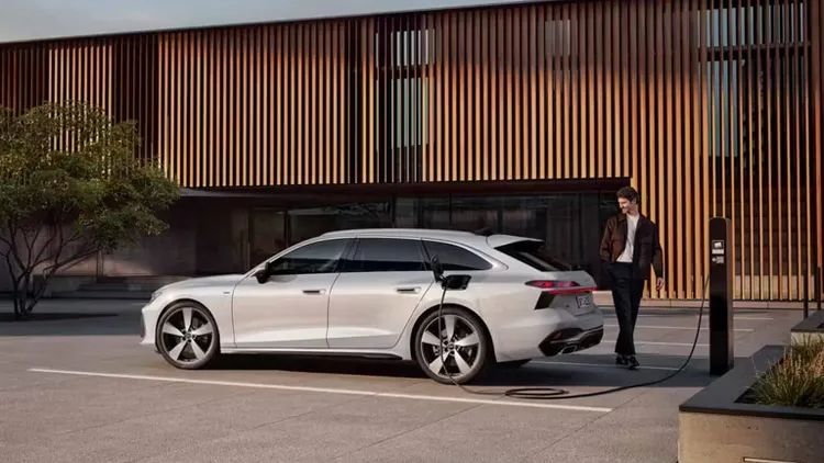 Muž prechádza okolo vozidla a pozerá sa neho. Vozidlo je zaparkované pred modernou budovou. Nabíjacia stanica je umiestnená v blízkosti vozidla. Elektrický automobil má elegantný dizajn a je bielej farby.