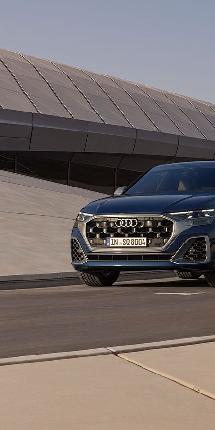 Audi SQ8 SUV je zaparkované pred modernou budovou. Žena vo svetlom overale prechádza okolo vozidla. Scéna je nasnímaná za denného svetla.