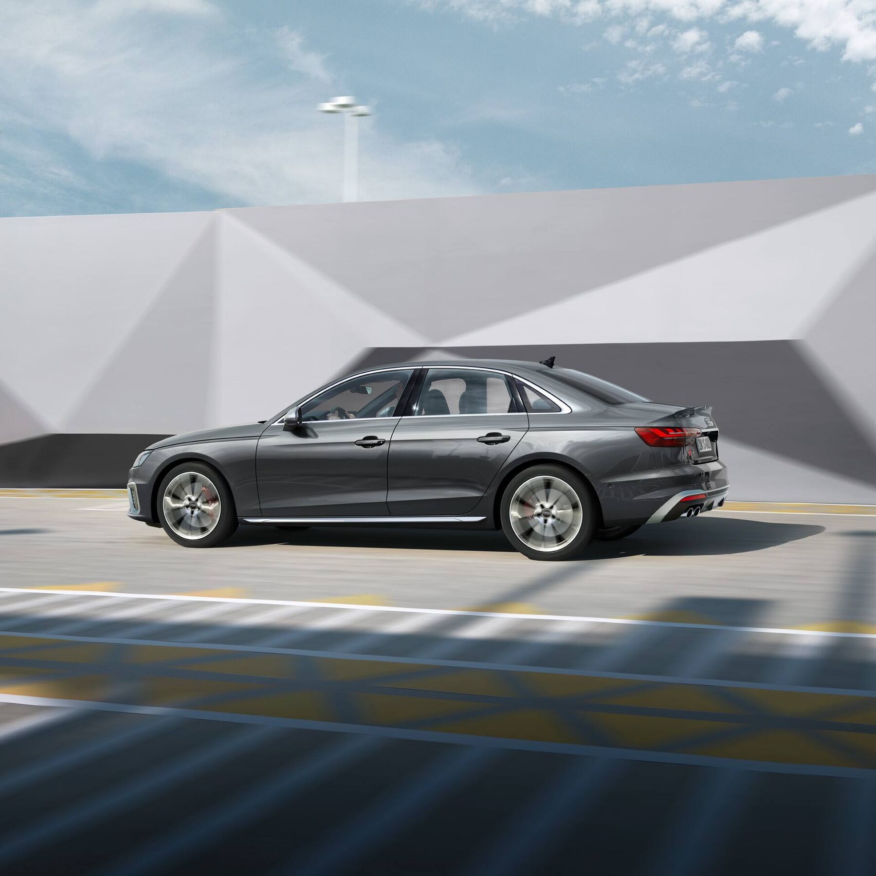 Moderné šedé Audi S4 Limuzína na ceste prechádza okolo zaujímavej budovy s geometrickými tvarmi. Auto je v pohybe a jeho dizajn je elegantný. Na oblohe sú jemné mraky.