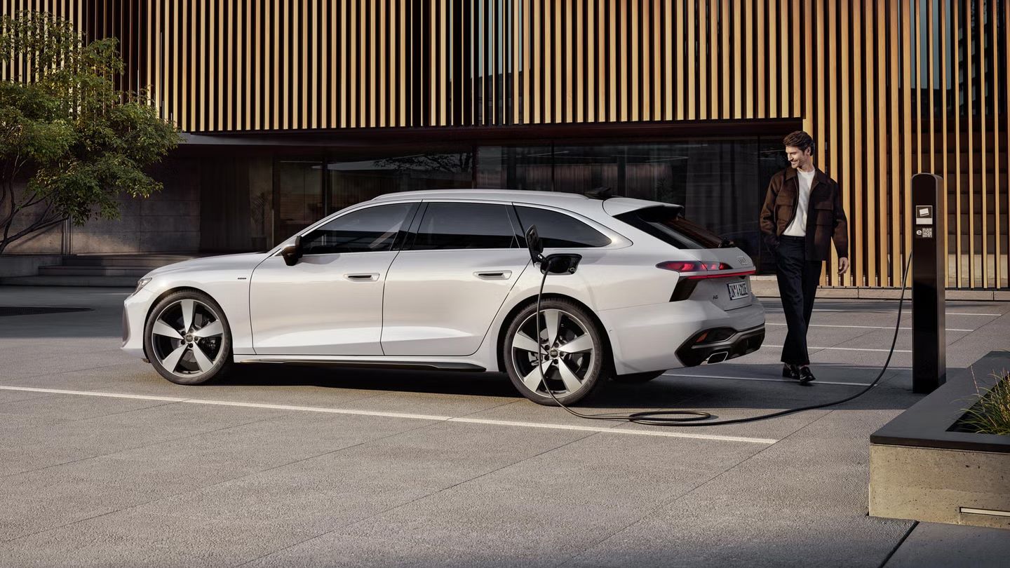 A6 Avant e-hybrid stojí bokom pred vchodom do veľkej budovy a nabíja sa pomocou napájacieho kábla. Za vozidlom stojí muž stredného veku.